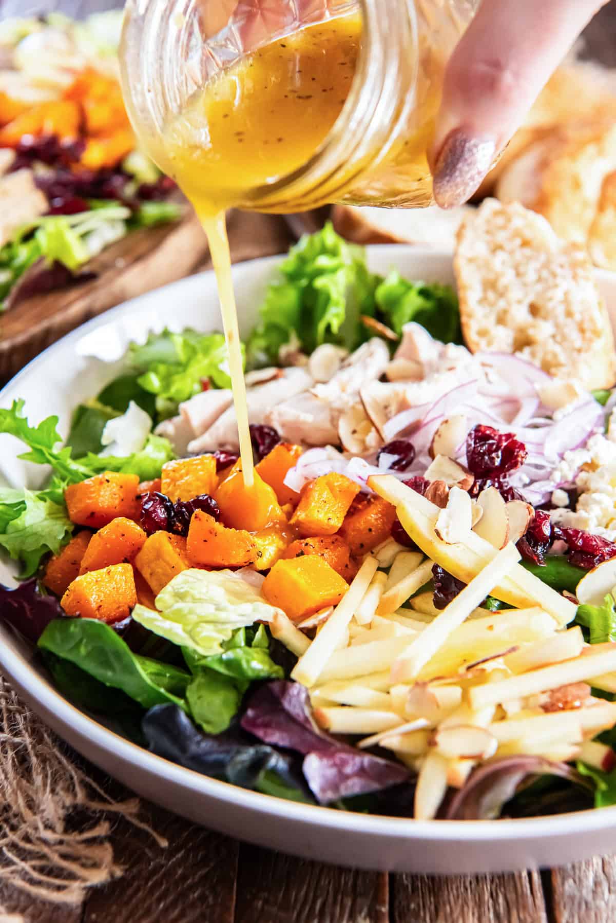 A hand pours maple vinaigrette over a bowl of butternut squash salad with chicken and apples.