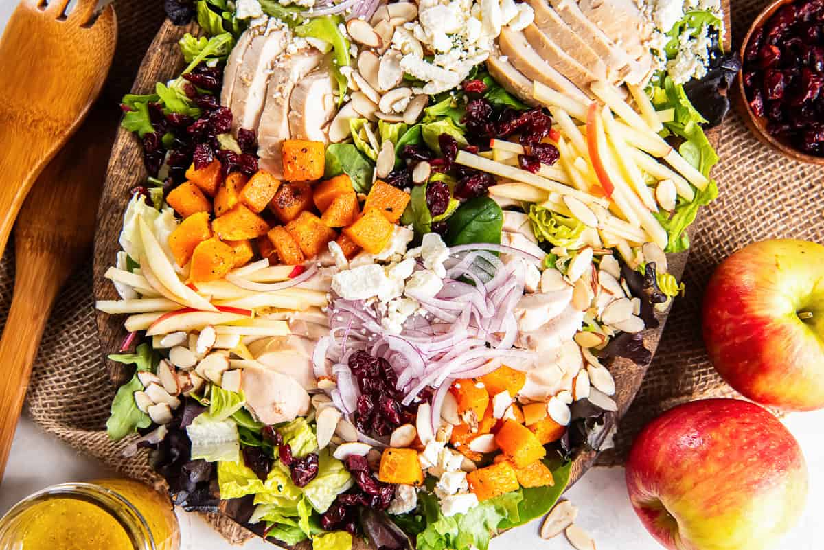 A large wooden platter filled with mixed greens, sliced chicken, roasted butternut squash, thin apple strips, red onion, dried cranberries, sliced almonds, and crumbled feta cheese, with fresh apples and maple vinaigrette alongside.