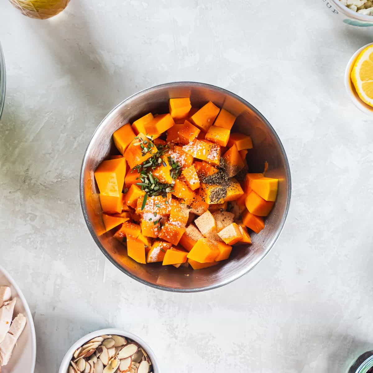 Cubed butternut squash with olive oil, rosemary, granulated garlic, salt, and pepper in a mixing bowl.