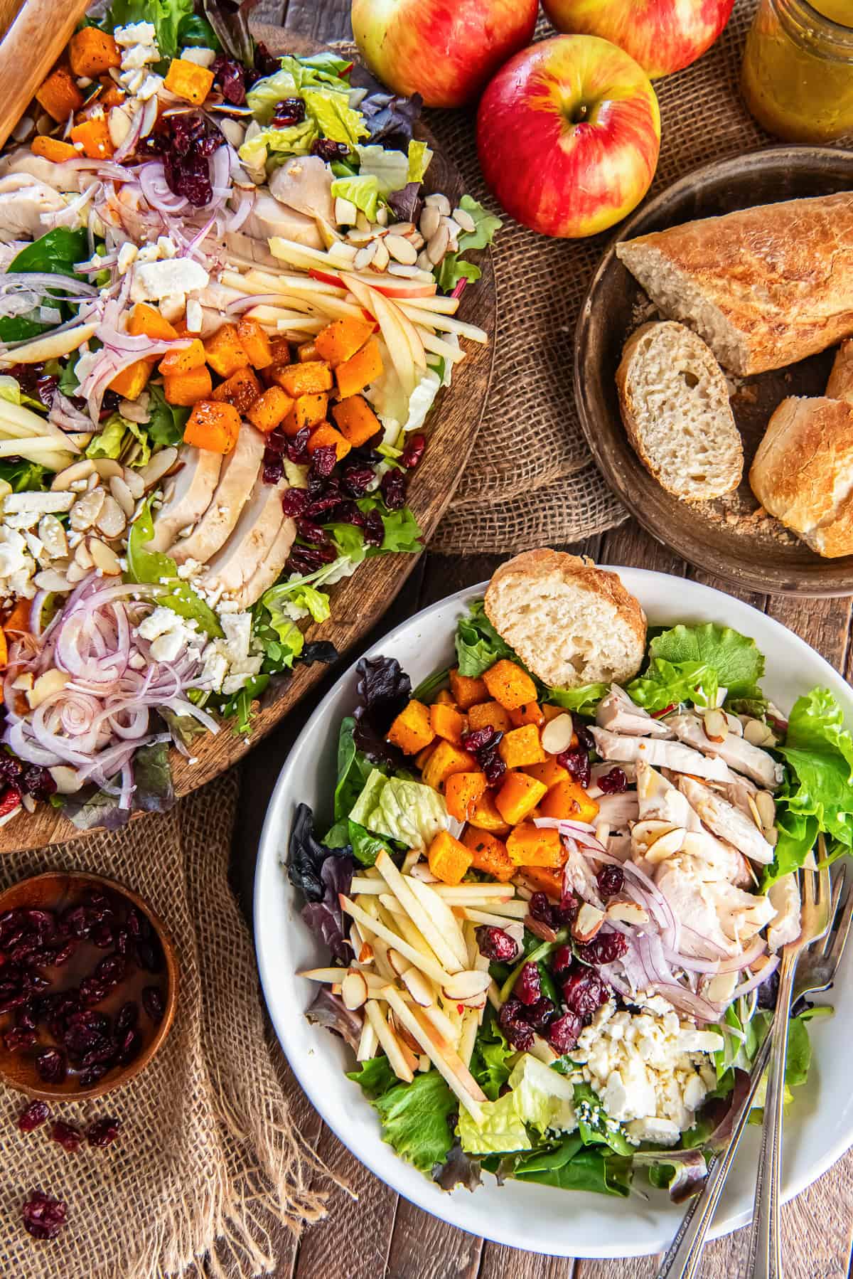 A white bowl filled with chicken salad, roasted butternut squash, sliced apples, cranberries, feta, and mixed greens sits beside a large wooden platter of salad. Fresh apples, crusty bread, and a jar of dressing complete the rustic table setting.