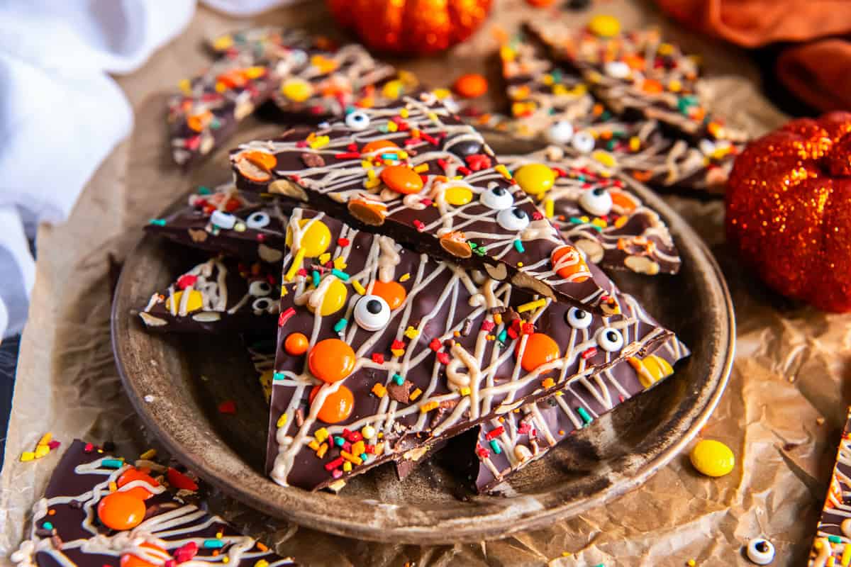 Halloween chocolate bark stacked on a rustic plate with peanuts visible along the edges, topped with white chocolate drizzle, Reese’s Pieces, sprinkles, and candy eyeballs.