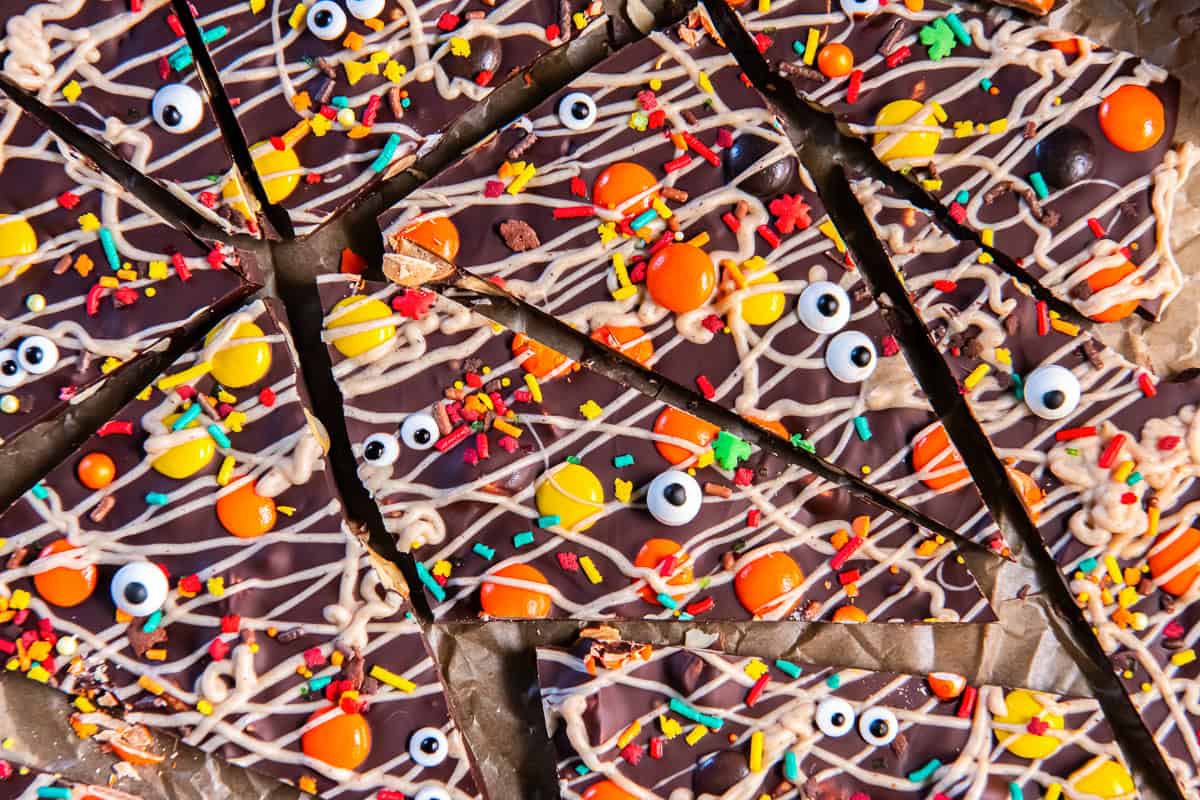 Halloween chocolate bark pieces on parchment, topped with white chocolate drizzle, Reese’s Pieces, colorful sprinkles, and candy eyeballs.