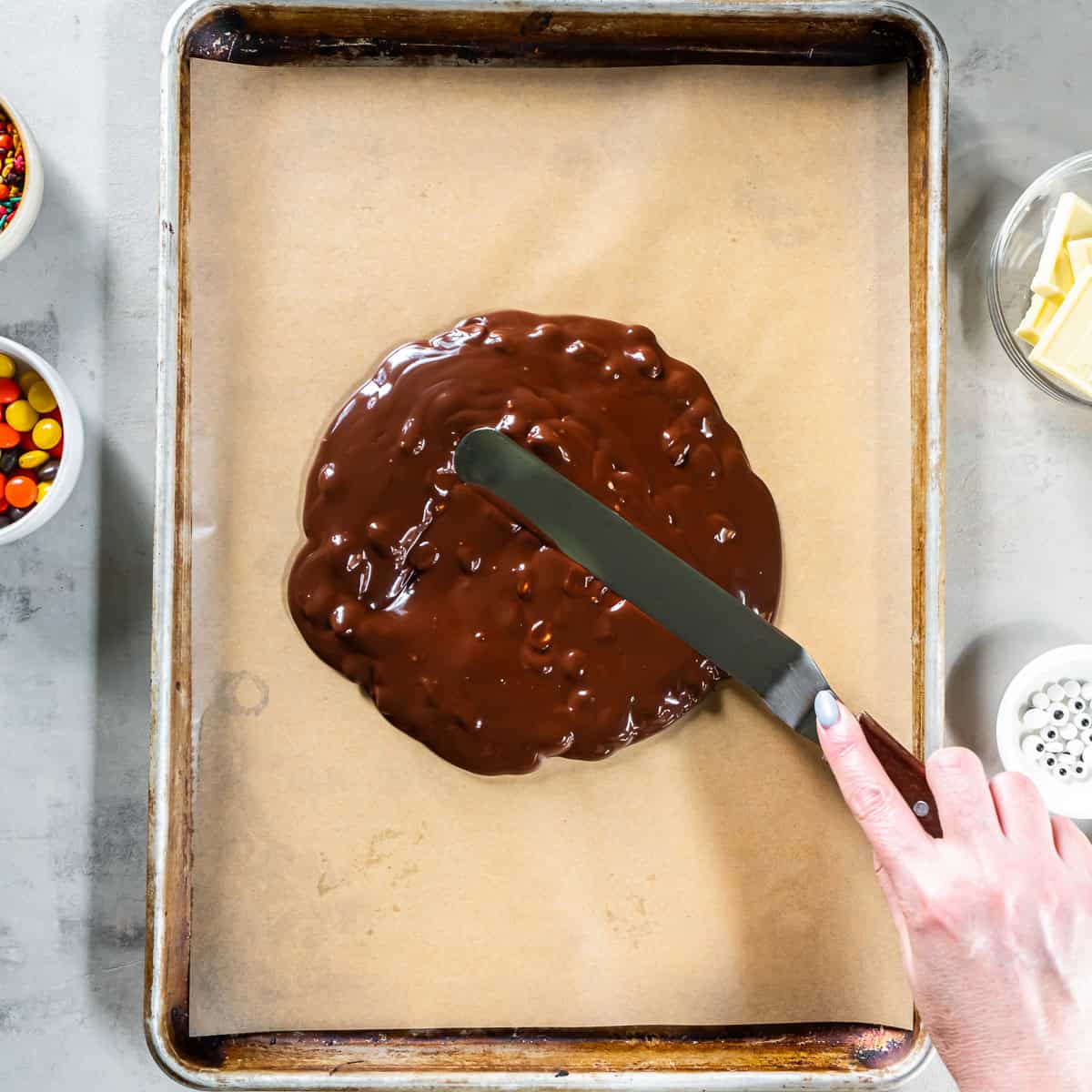 Spreading melted chocolate and peanuts onto a parchment-lined baking sheet with an offset spatula.