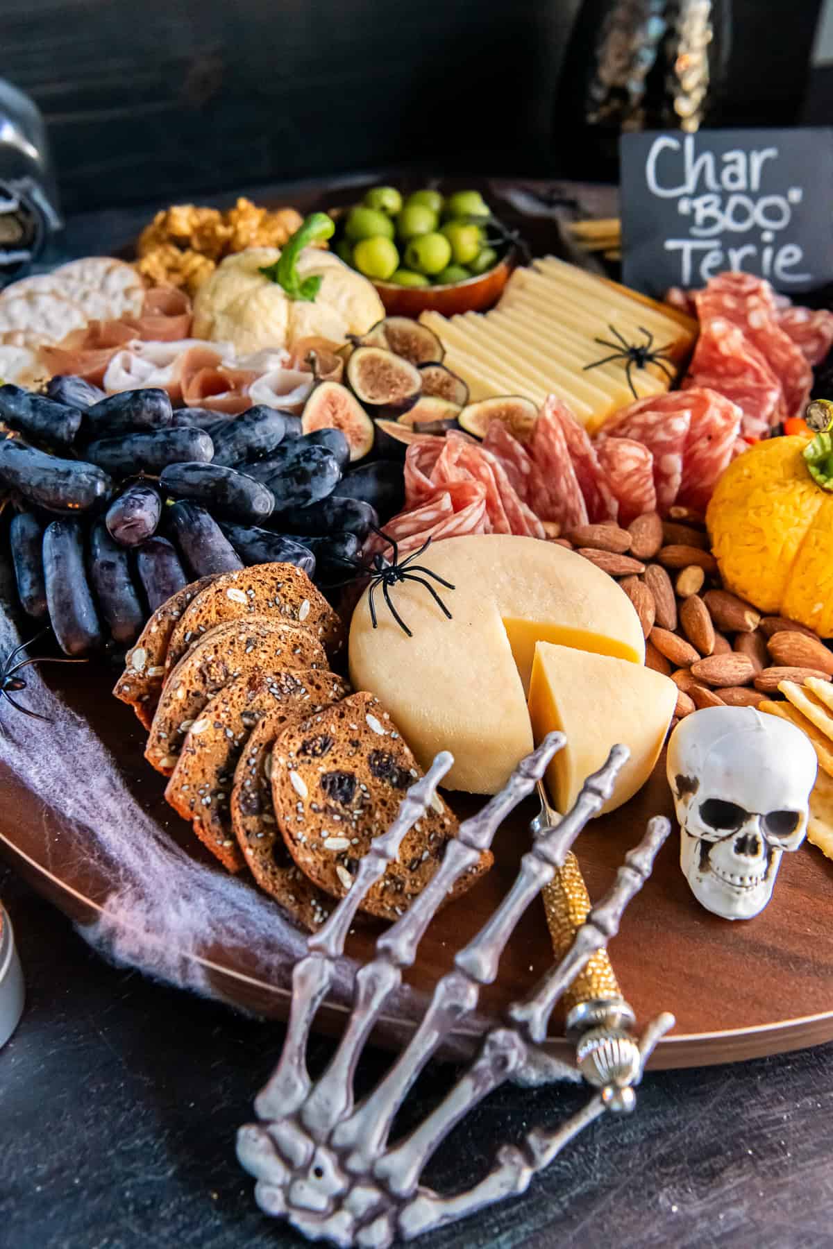 A skeleton hand resting on the edge of a Halloween charcuterie board with salami, sliced cheese, grapes, crackers, and a plastic skull.