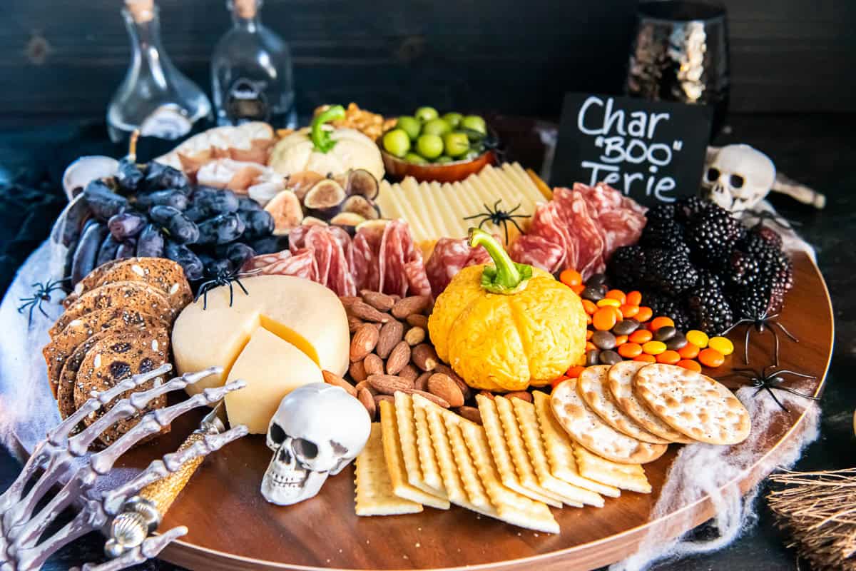 Halloween charcuterie board with orange and white Boursin cheese pumpkins, sliced cheeses, cured meats, grapes, blackberries, figs, olives, almonds, crackers, and colorful candies, decorated with skeleton hands, skulls, spiders, and a “Char BOO Terie” sign.
