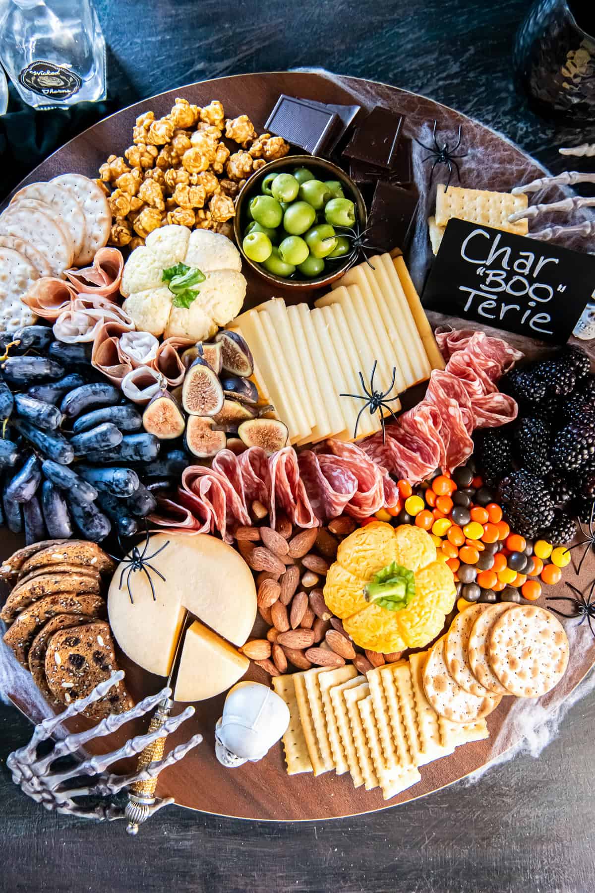 Halloween charcuterie board with two pumpkin-shaped cheese balls, sliced cheeses, folded salami and prosciutto, grapes, figs, blackberries, olives, crackers, almonds, caramel popcorn, chocolate, and colorful candies, decorated with spiders, cobwebs, and a skeleton hand.