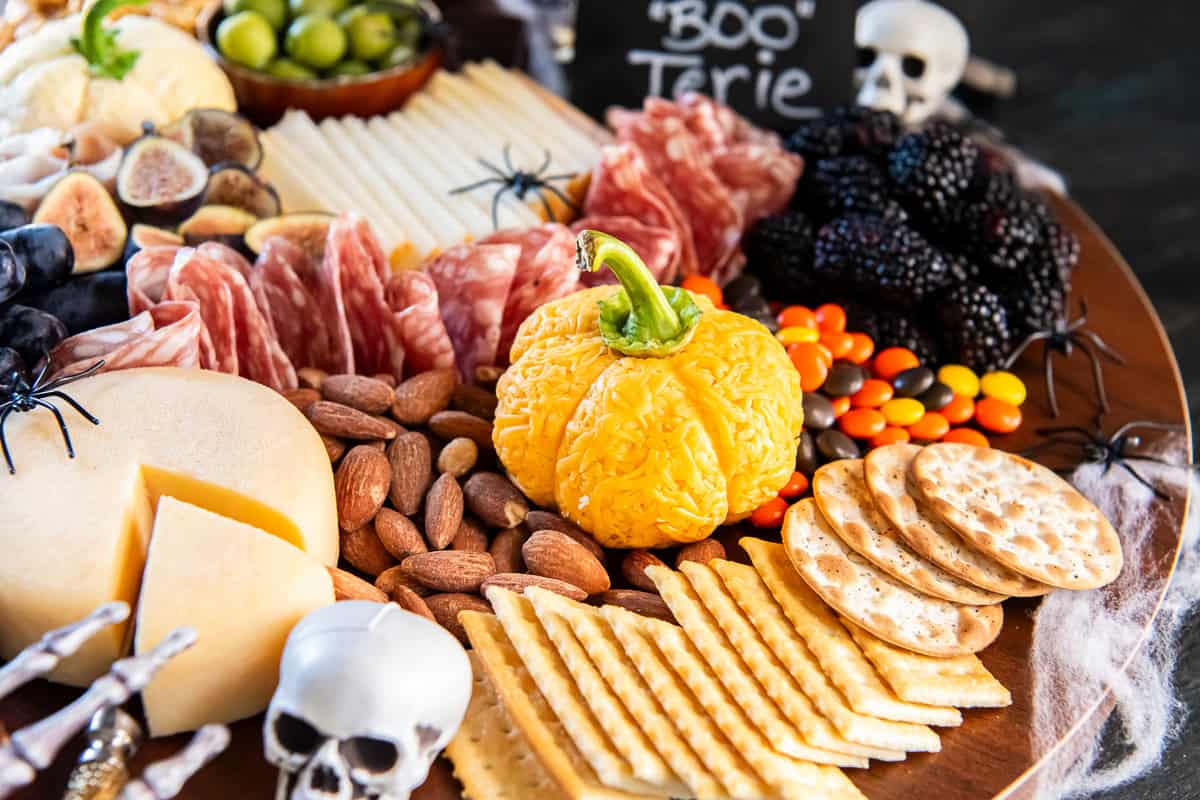 : Orange pumpkin-shaped Boursin cheese ball with a green bell pepper stem, nestled among almonds, salami, blackberries, colorful candies, and crackers on a Halloween charcuterie board.