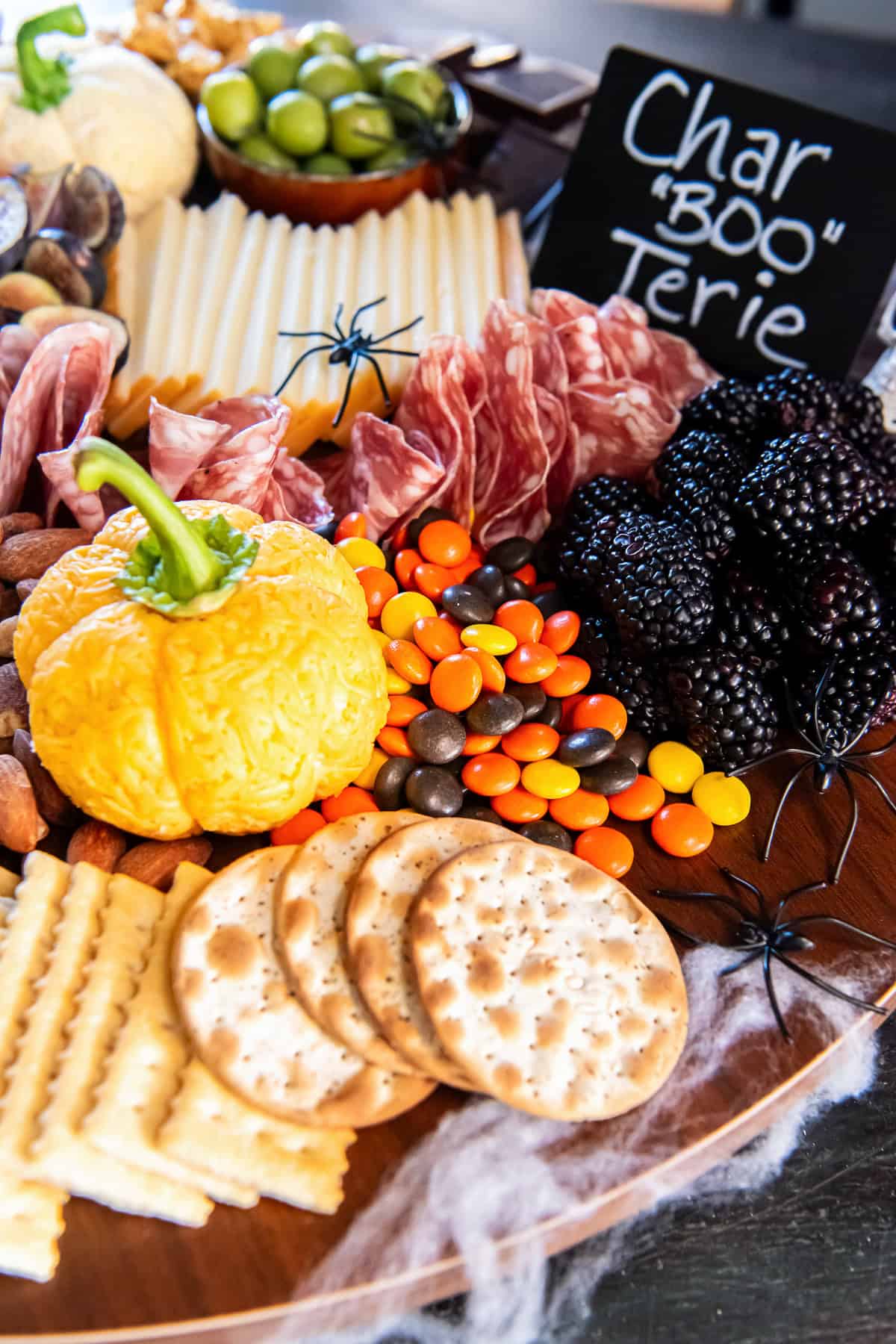 Orange pumpkin-shaped Boursin cheese ball surrounded by folded salami, sliced cheese, blackberries, almonds, colorful candies, and crackers on a Halloween charcuterie board decorated with black spiders and a “Char BOO Terie” sign.