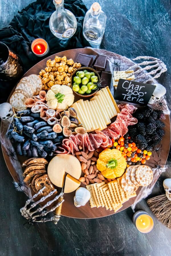 Halloween charcuterie board filled with pumpkin-shaped cheese balls, sliced cheeses, cured meats, grapes, figs, blackberries, olives, crackers, almonds, popcorn, chocolate, and candy, surrounded by cobwebs, spiders, skulls, candles, and skeleton hands.
