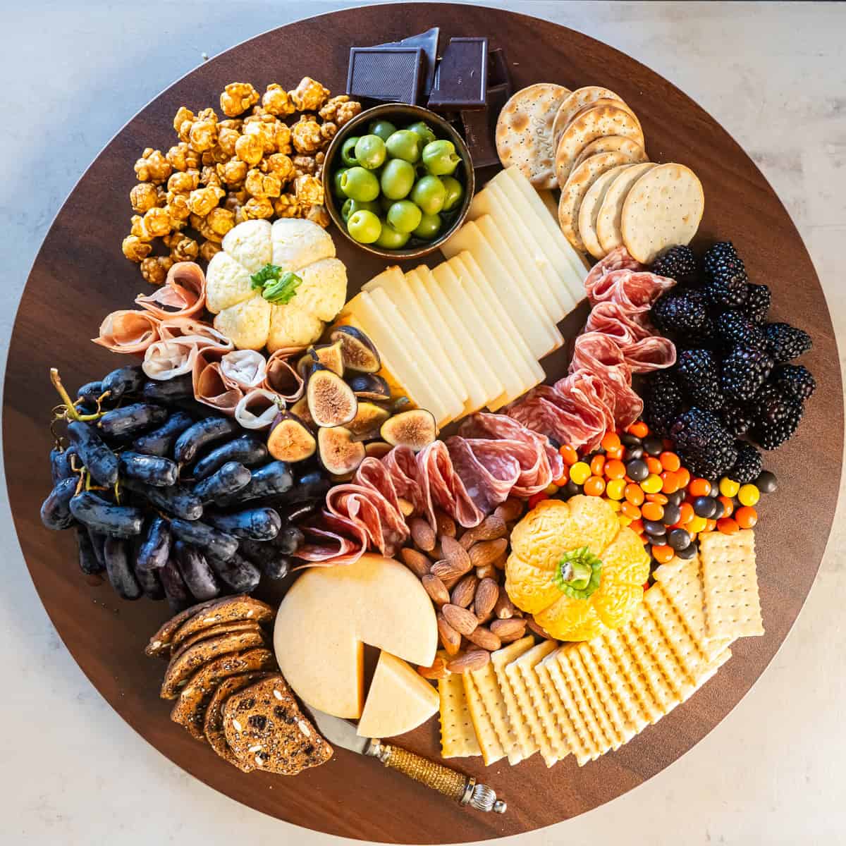A fully assembled Halloween charcuterie board with two cheese pumpkins, cured meats, assorted cheeses, grapes, blackberries, figs, crackers, olives, almonds, caramel popcorn, dark chocolate, and colorful candy.