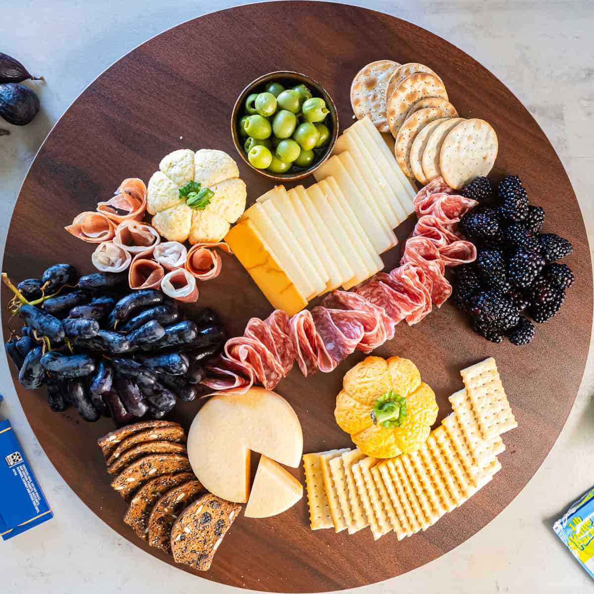 A partially assembled charcuterie board with Boursin cheese pumpkins, cured meats, sliced cheeses, dark grapes, blackberries, assorted crackers, and green olives.