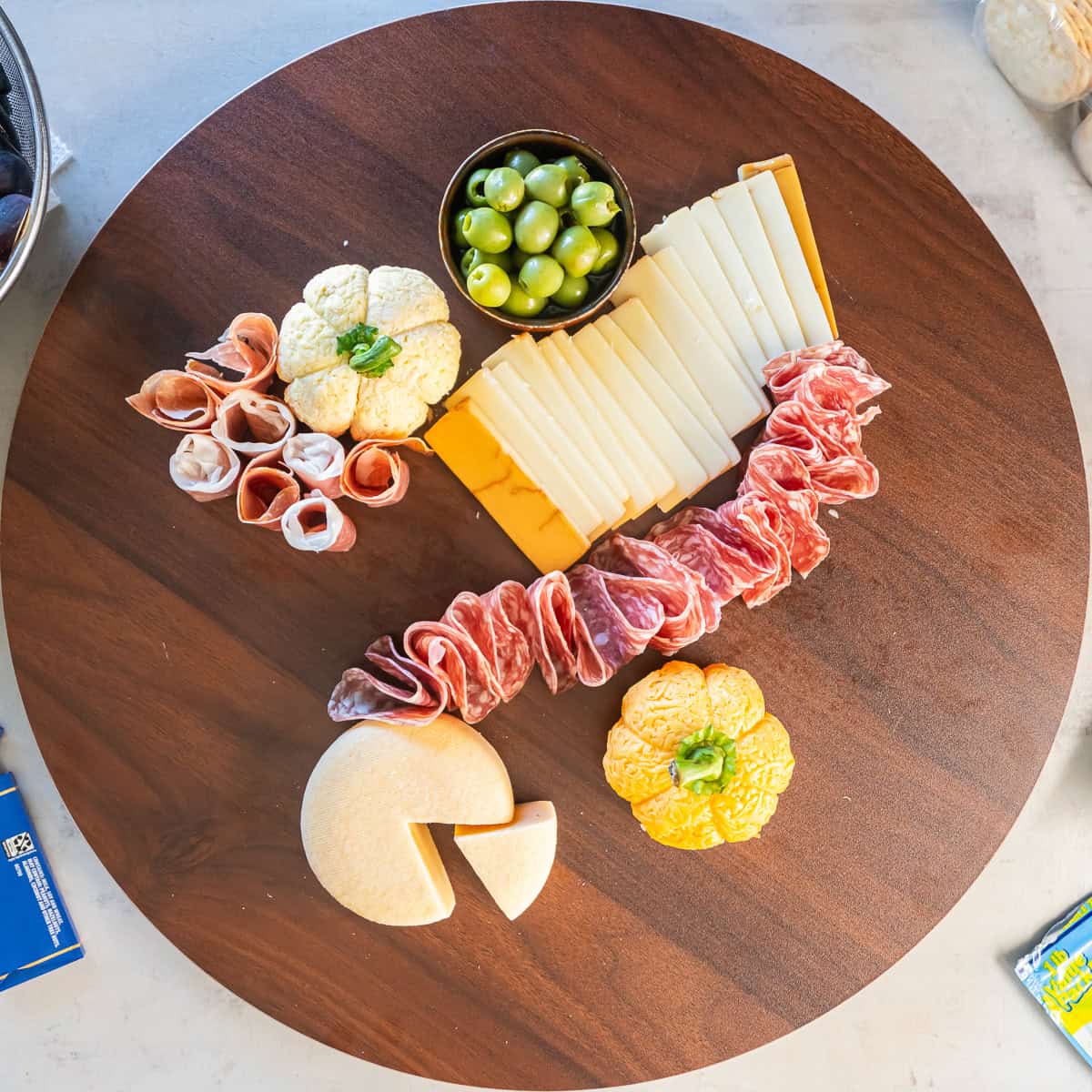 A round wooden charcuterie board with folded salami and prosciutto arranged around sliced cheeses, two Boursin cheese pumpkins, a Gouda wedge, and a bowl of green olives.