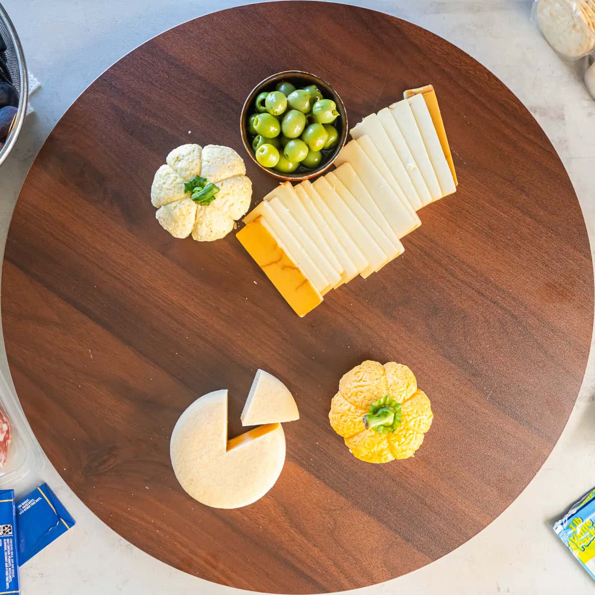A round wooden charcuterie board with two Boursin cheese pumpkins, sliced cheeses, a wedge of Gouda, and a small bowl of green olives arranged with space between them.