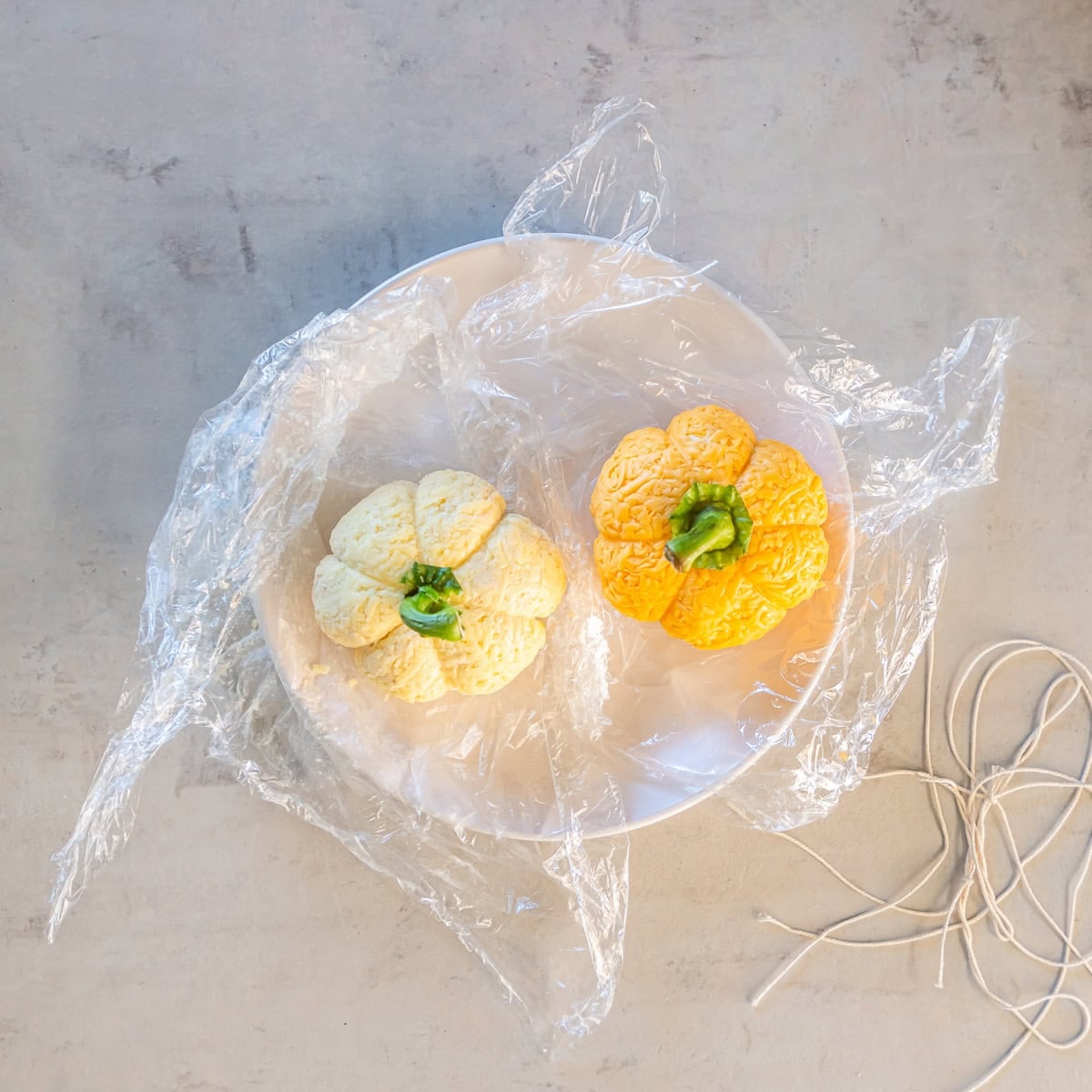 Two finished Boursin cheese pumpkins, one coated in white cheese and the other in orange cheddar, topped with green bell pepper stems on a white plate lined with plastic wrap.
