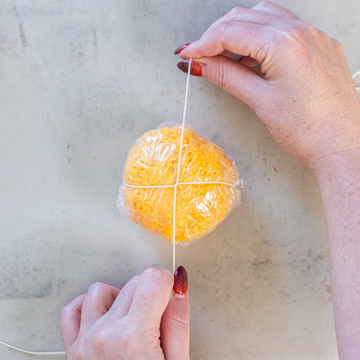 Hands pulling kitchen twine vertically around a plastic-wrapped, cheddar-coated Boursin cheese ball to shape it into a pumpkin.