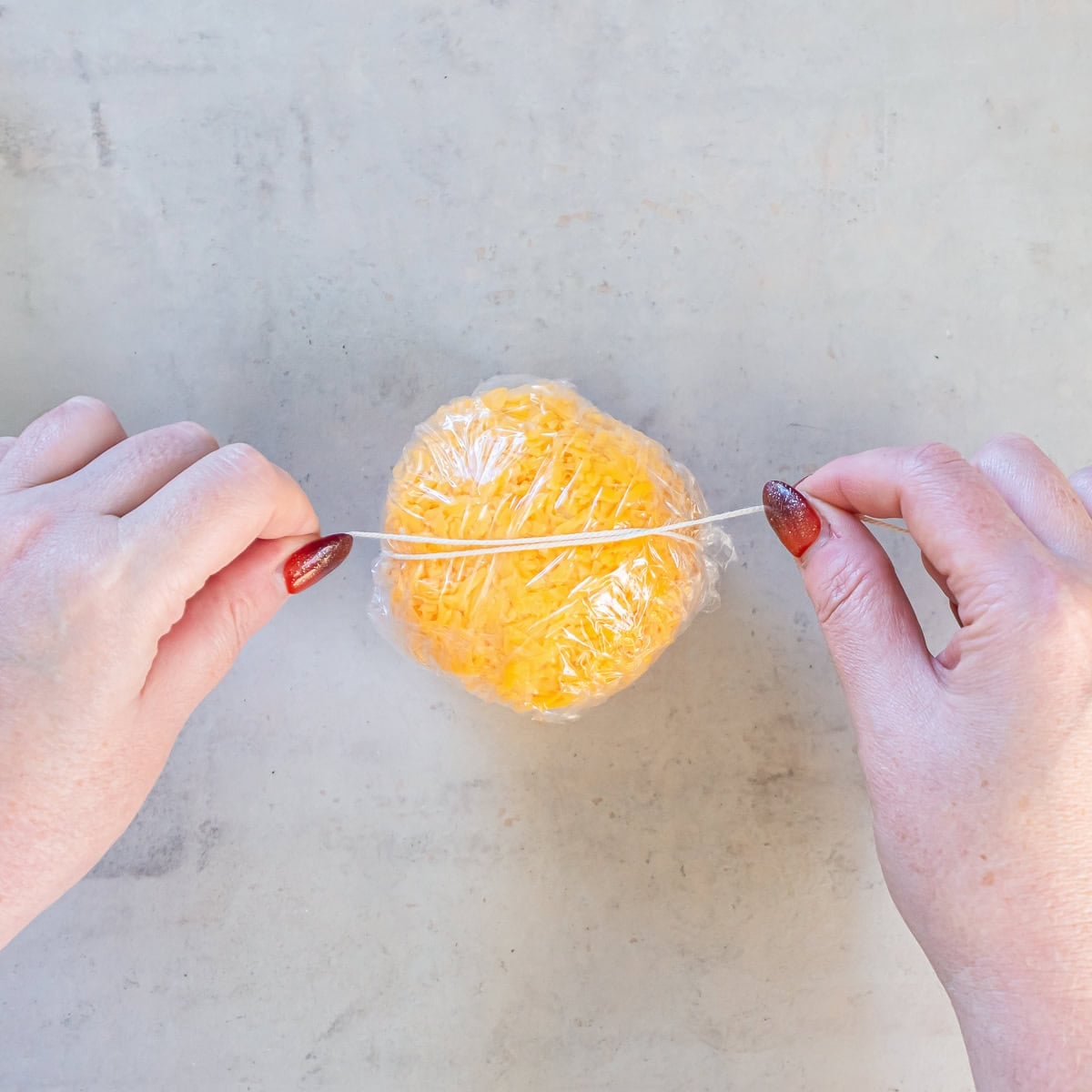 Hands pulling kitchen twine horizontally around a plastic-wrapped, cheddar-coated Boursin cheese ball.