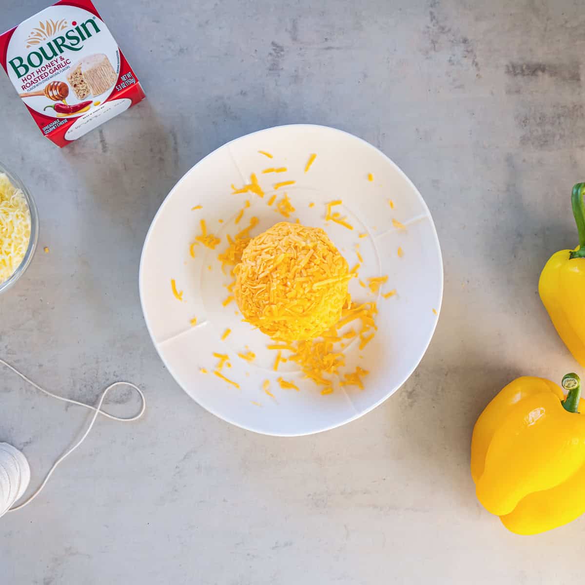 Round Boursin cheese ball coated in shredded orange cheddar on a white plate, with a Boursin package, shredded cheese, kitchen twine, and yellow bell peppers nearby.