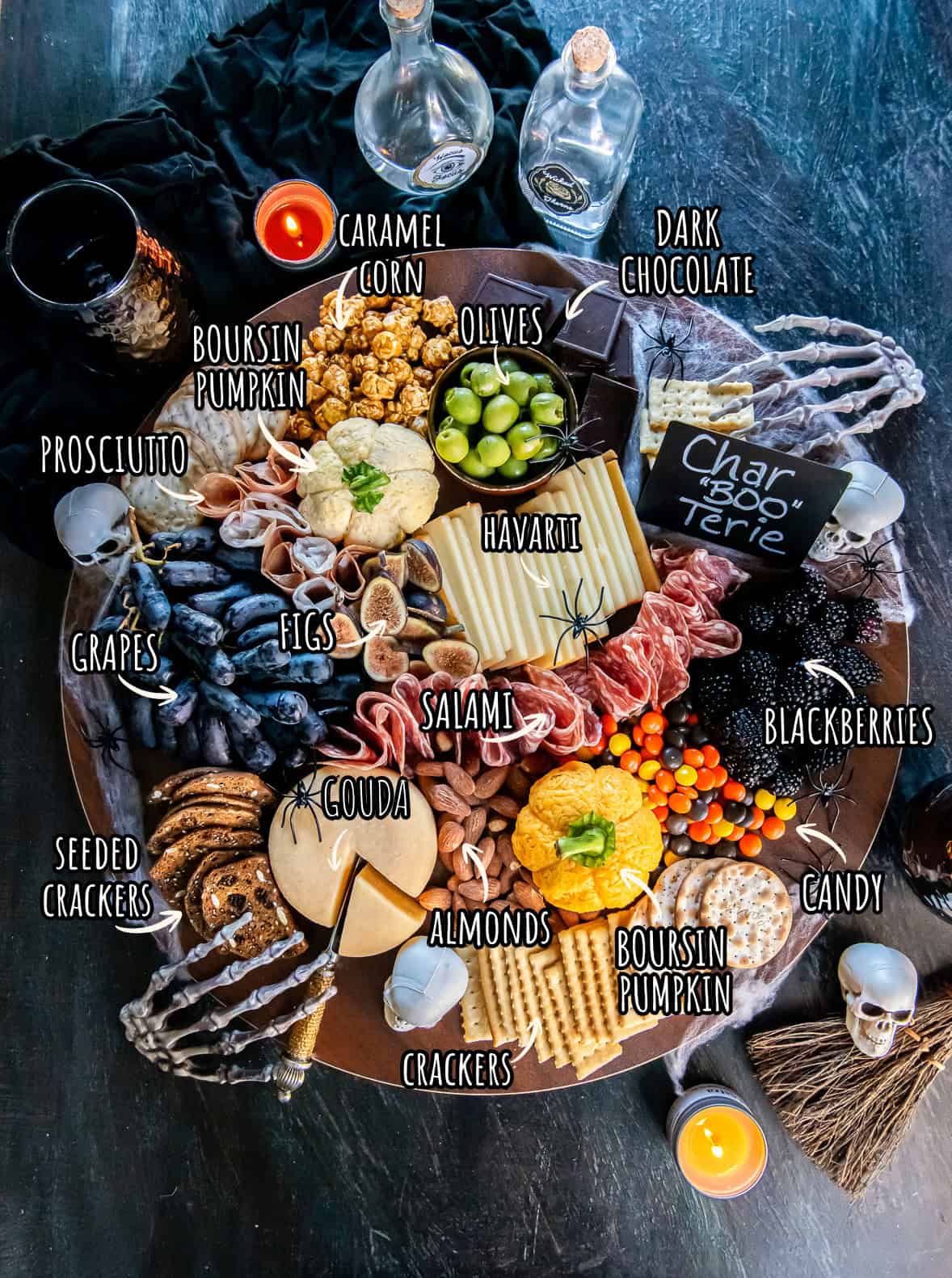 Ingredients for a Halloween charcuterie board arranged on a platter, with white labels and orange arrows identifying Boursin cheese pumpkins, meats, cheeses, fruit, crackers, nuts, olives, chocolate, and candy.