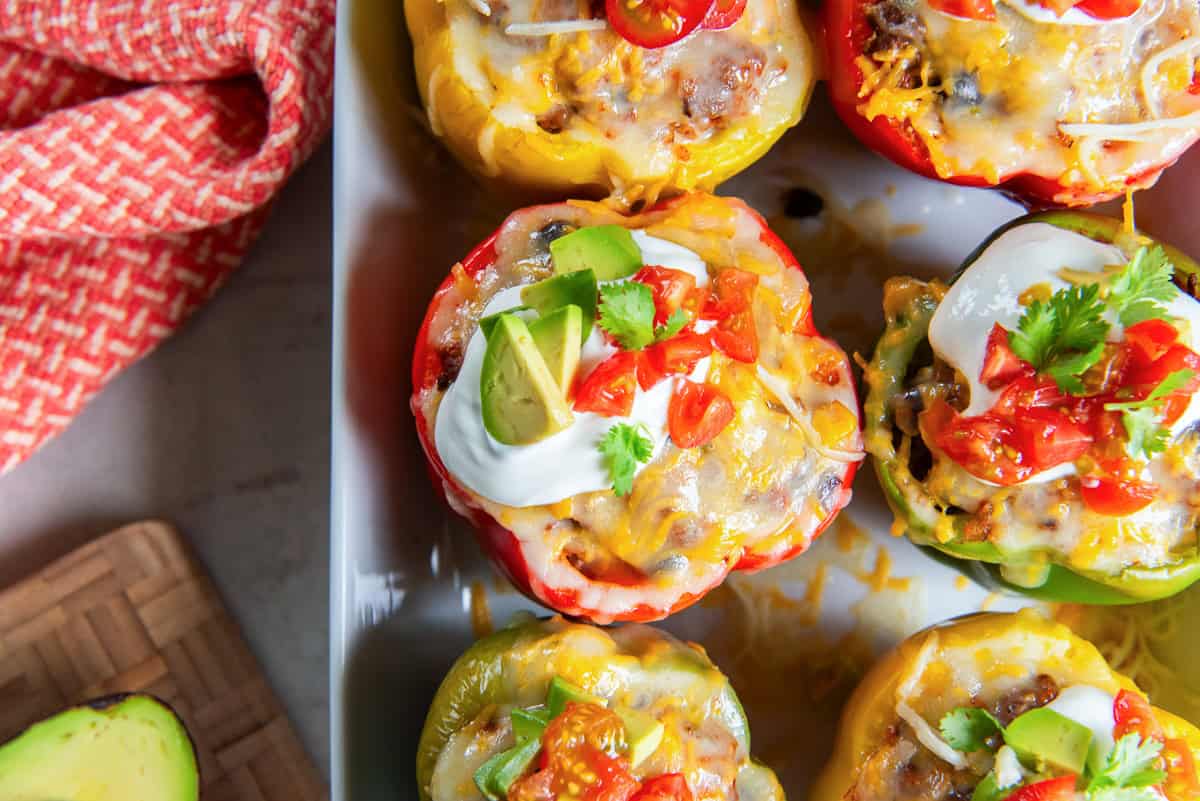 Mexican stuffed peppers topped with melted cheese, sour cream, avocado, diced tomatoes, and fresh cilantro in a white baking dish.