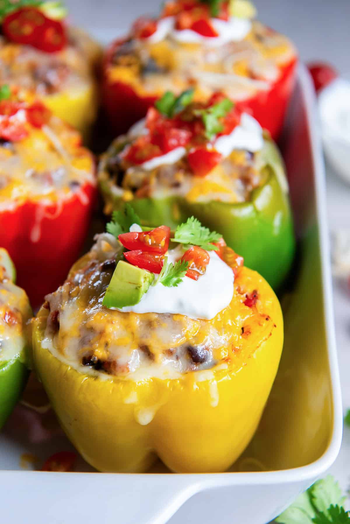 Close-up of a yellow Mexican stuffed pepper topped with melted cheese, sour cream, avocado, diced tomatoes, and cilantro in a baking dish.
