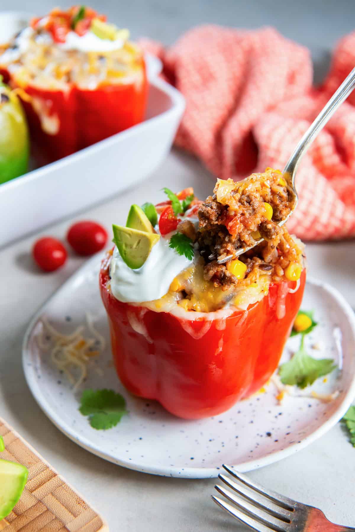 A fork breaking into a Mexican stuffed bell pepper topped with melted cheese, sour cream, avocado, diced tomatoes, and cilantro on a white plate.