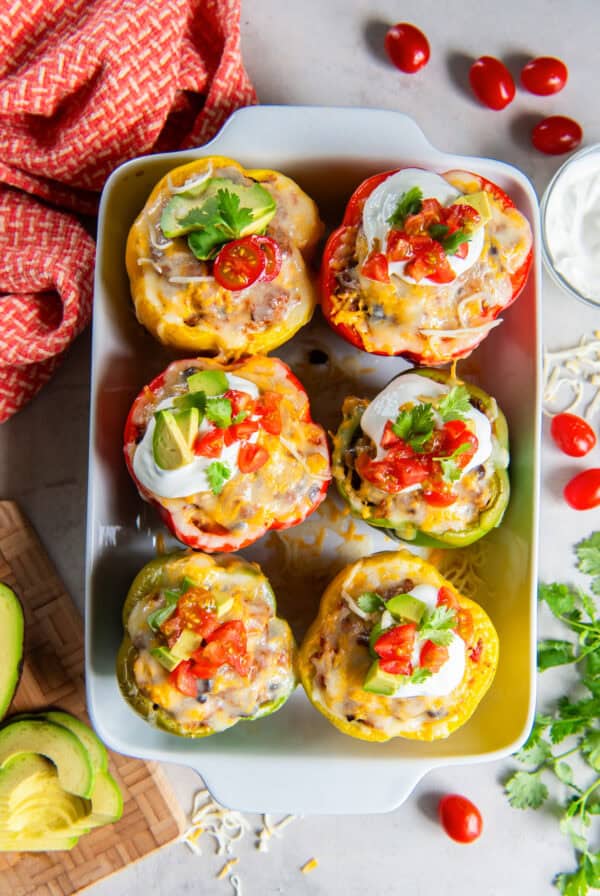 Mexican Stuffed Peppers topped with melted cheese, sour cream, avocado, diced tomatoes, and cilantro in a white baking dish.