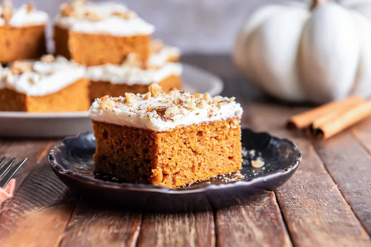 A single pumpkin bar with cream cheese icing on a black plate with a white pumpkin behind it.