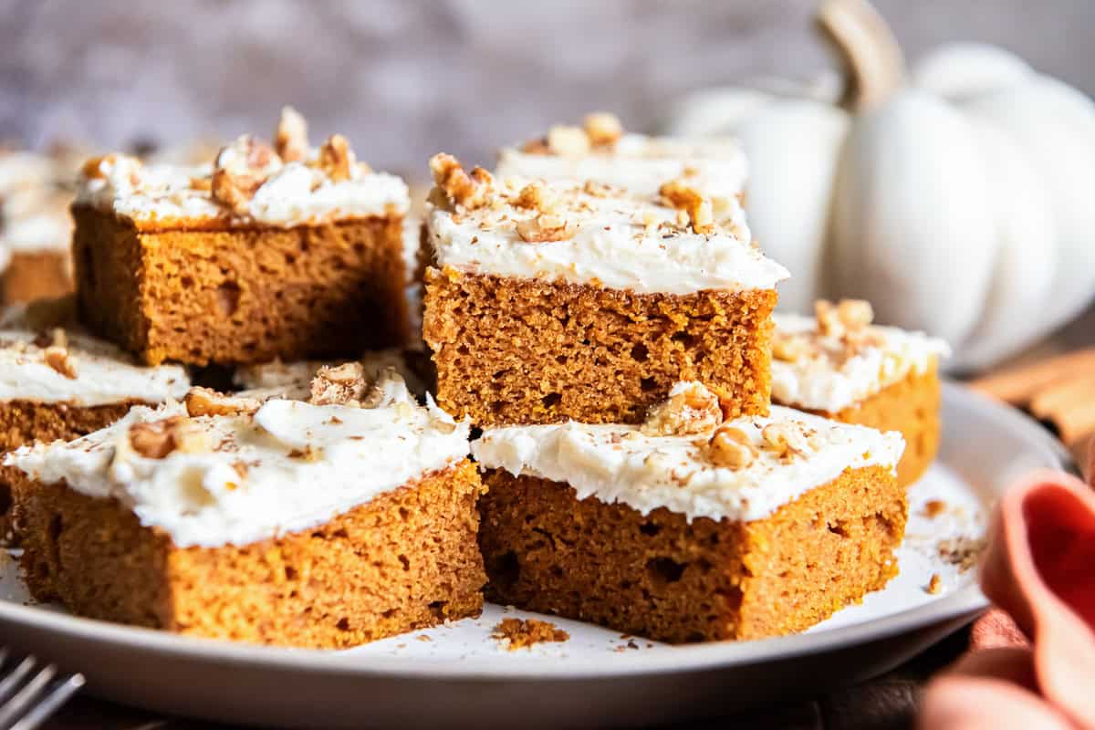 Pumpkin bars topped with cream cheese frosting, chopped walnuts, and cinnamon arranged on a serving platter with a white pumpkin in the background.