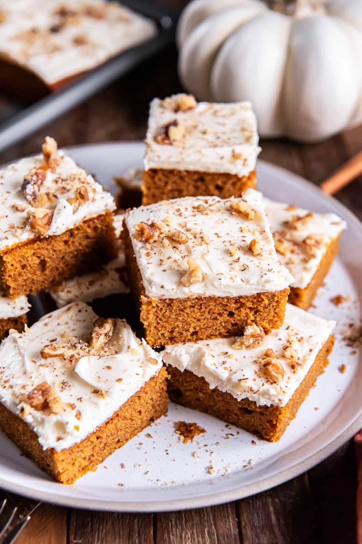 Pumpkin bars topped with cream cheese frosting, chopped walnuts, and cinnamon are stacked on a serving platter.