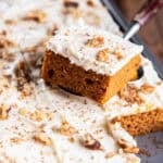A pumpkin bar topped with cream cheese frosting and chopped walnuts is lifted from a pan of frosted pumpkin bars.