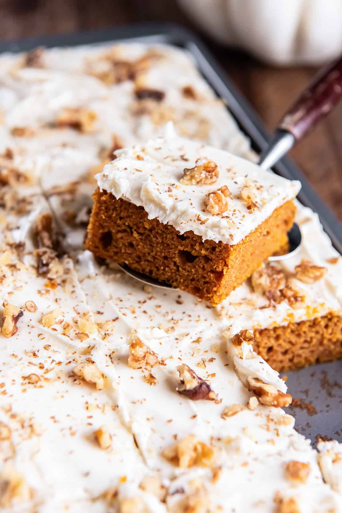 A pumpkin bar topped with cream cheese frosting and chopped walnuts is lifted from a pan of frosted pumpkin bars.