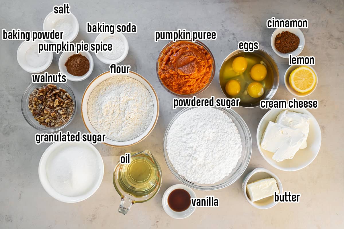 Ingredients for pumpkin bars with cream cheese frosting arranged in bowls, including pumpkin puree, flour, sugars, eggs, oil, spices, cream cheese, butter, lemon, vanilla, and walnuts.