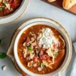 Two bowls of lasagna soup next to breadsticks on a board.