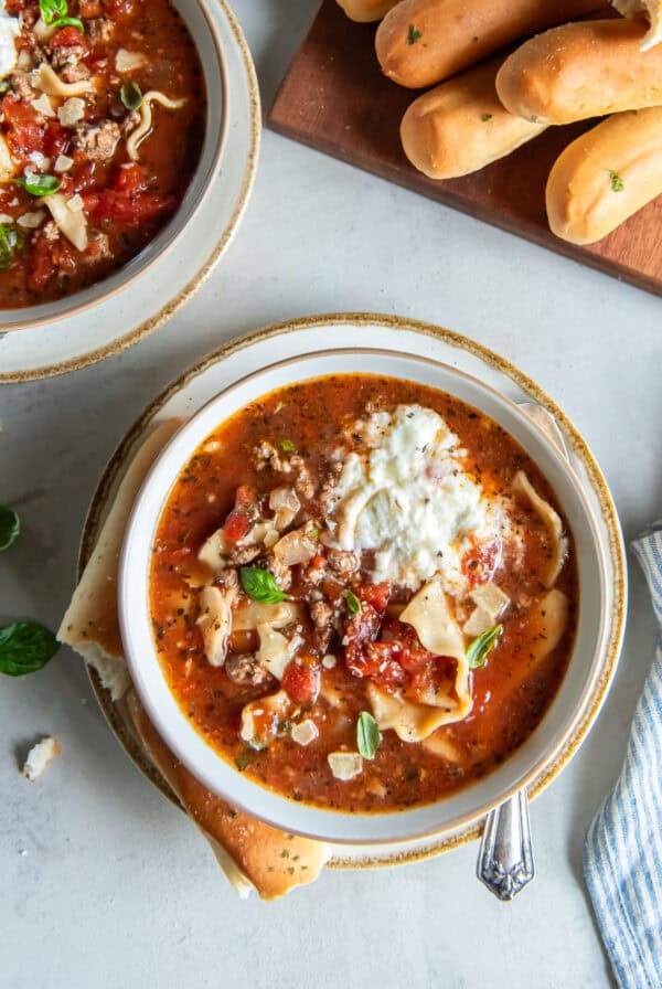 Two bowls of lasagna soup next to breadsticks on a board.