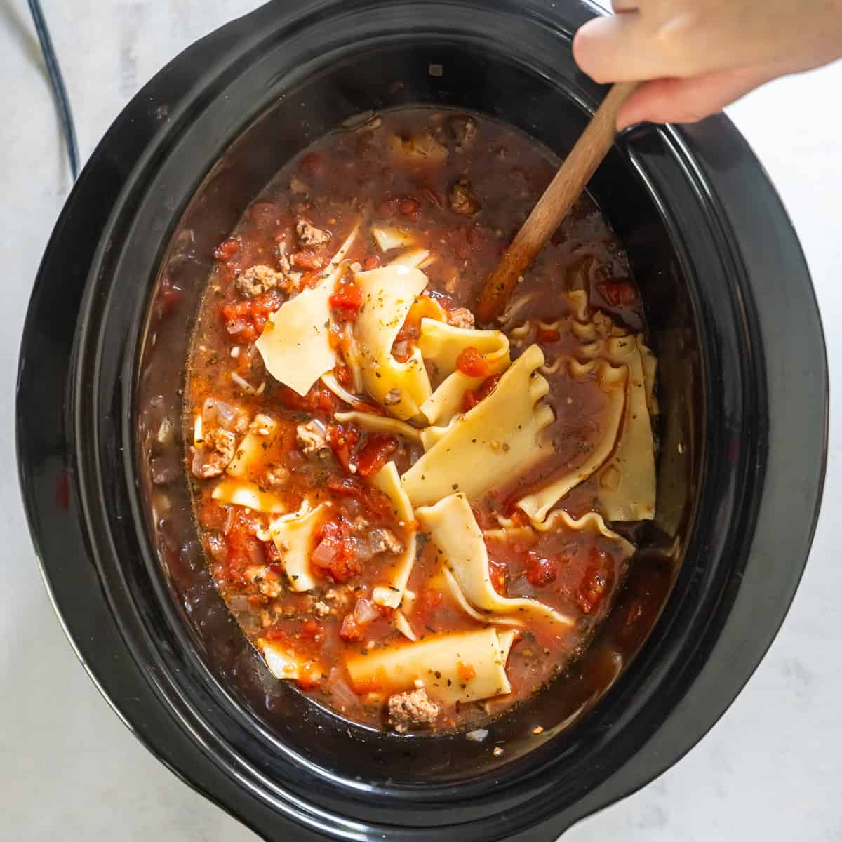 A wooden spoon stirring lasagna soup in a slow cooker.