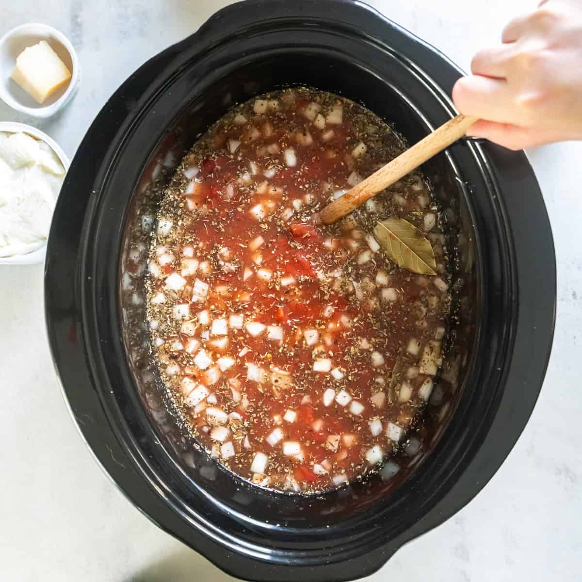 A hand holding a wooden spoon stirring the soup base for lasagna soup in a slow cooker.