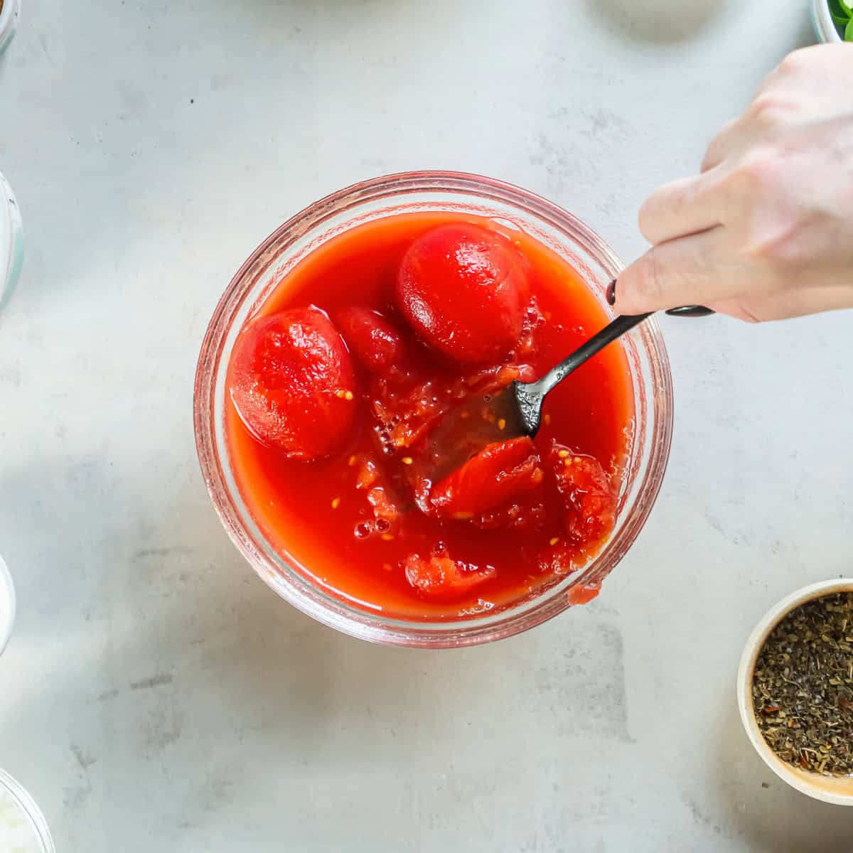 A fork breaking up whole San Marzano tomatoes in a small glass bowl.