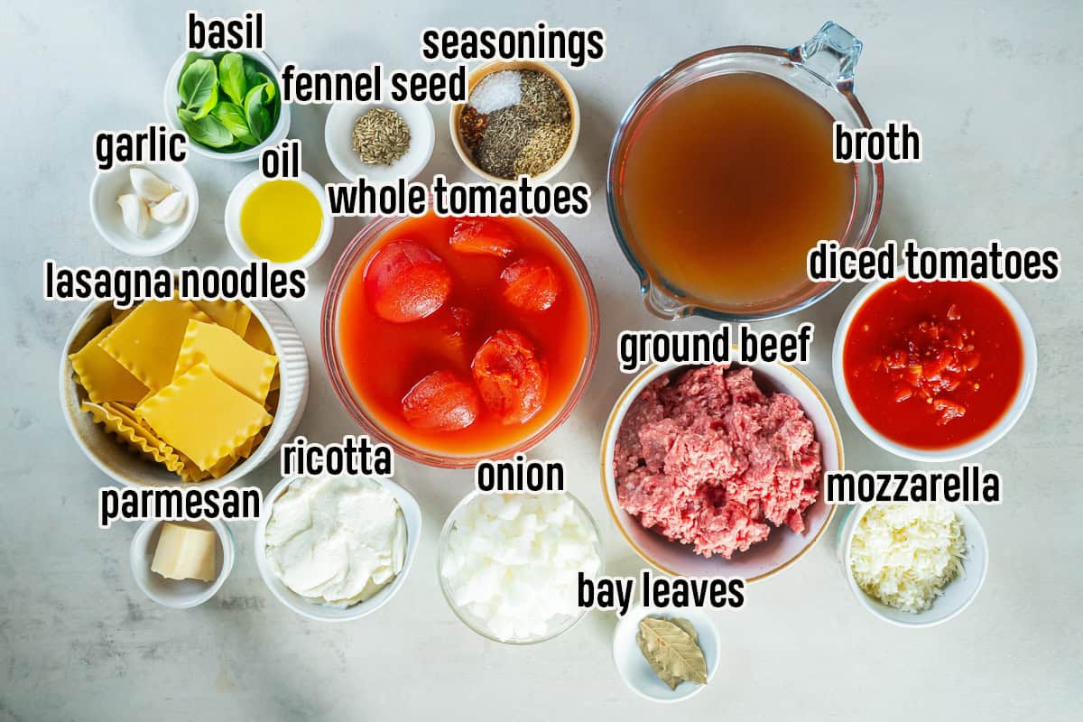 Slow cooker lasagna soup ingredients arranged in bowls, including ground beef, broken lasagna noodles, whole and diced tomatoes, broth, onion, garlic, olive oil, basil, fennel seed, seasonings, bay leaves, ricotta, mozzarella, and Parmesan.